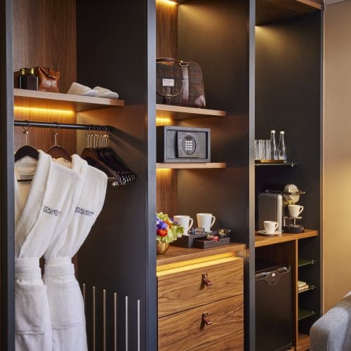 Modern hotel closet with in-room amenities including bathrobes, safe, coffee machine, and mini-fridge, all softly lit.