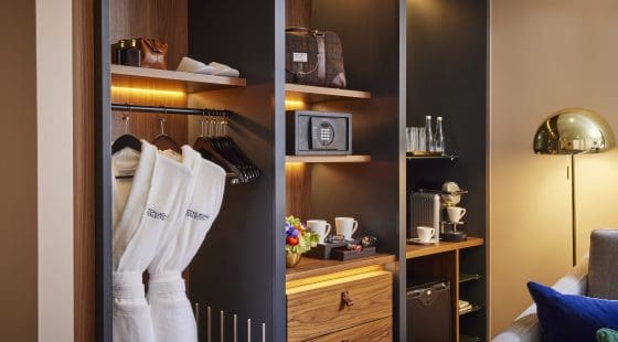 Modern hotel closet with in-room amenities including bathrobes, safe, coffee machine, and mini-fridge, all softly lit.