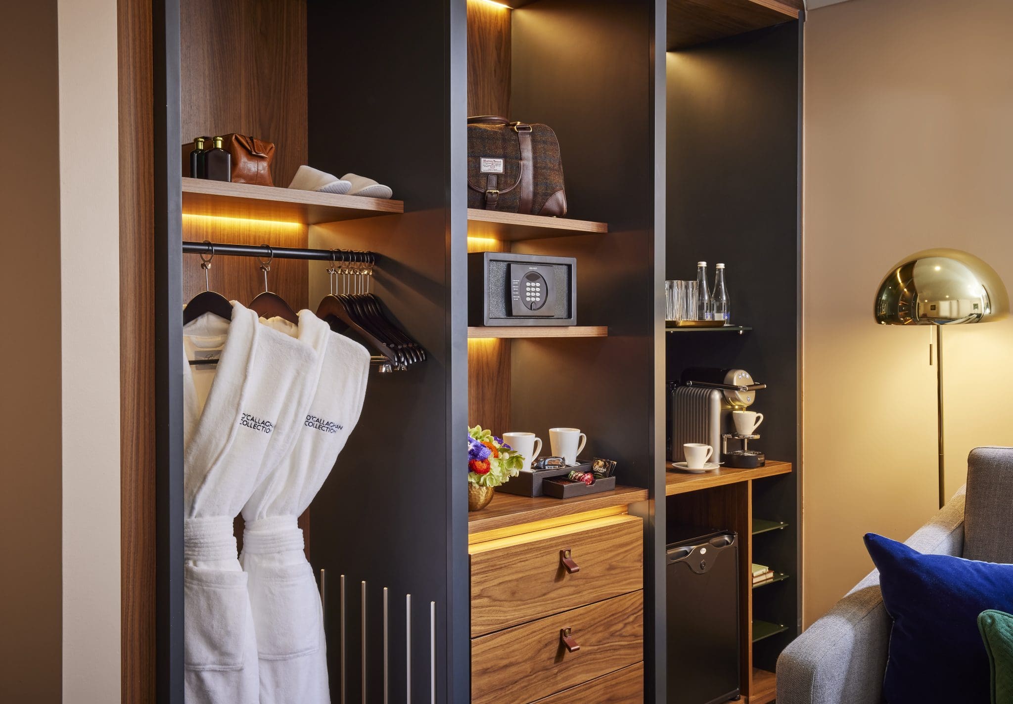 Modern hotel closet with in-room amenities including bathrobes, safe, coffee machine, and mini-fridge, all softly lit.
